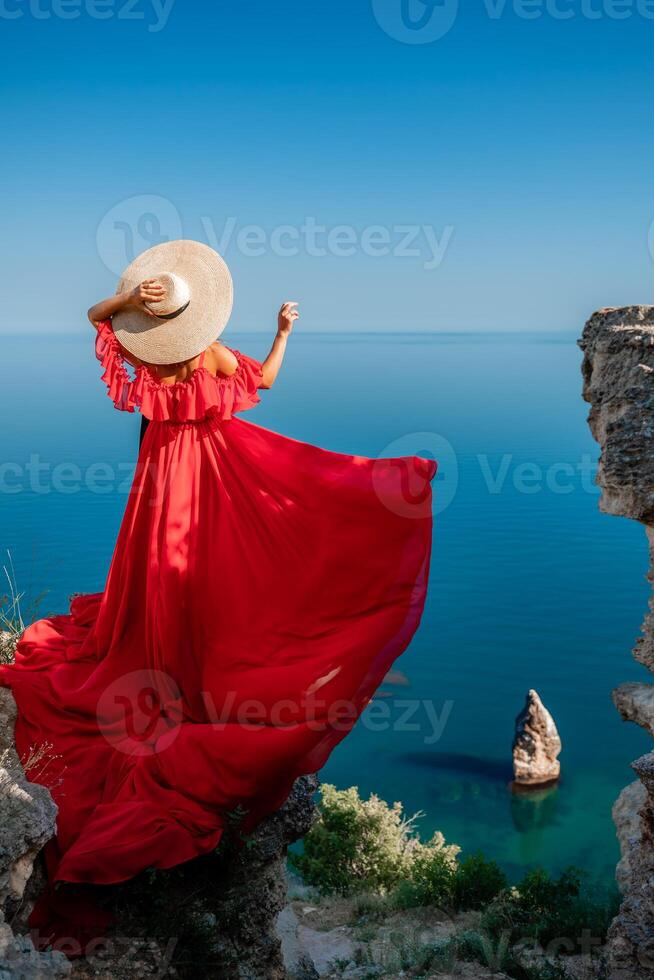 Red dress sea woman. Happy woman with flowing hair in a long flowing red dress stands on a rock near the sea. Travel concept, photo session at sea