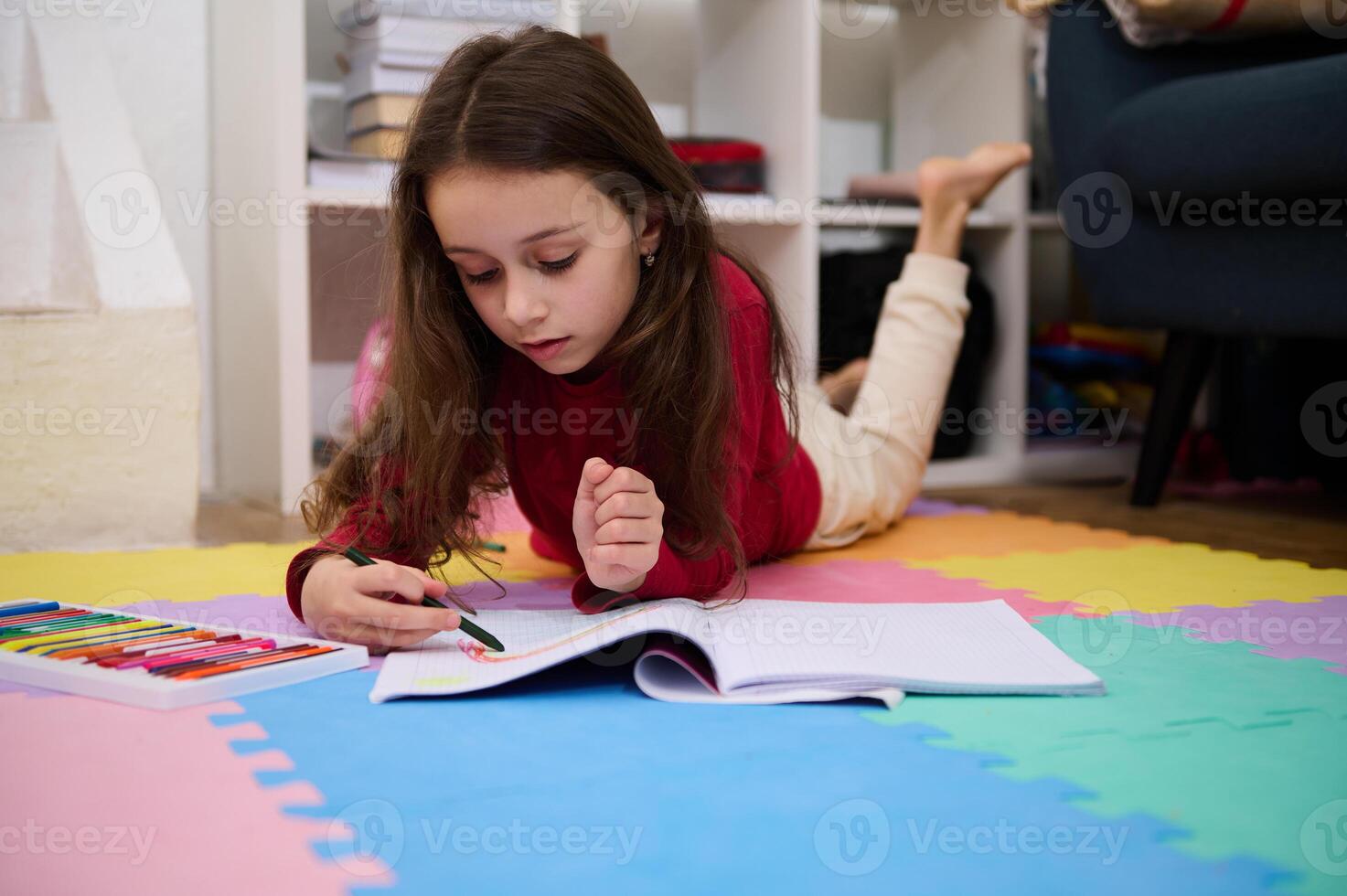 Authentic portrait of Caucasian elementary age school child girl doing homework, drawing picture with colorful, pastel pencils, writing and reading at home photo