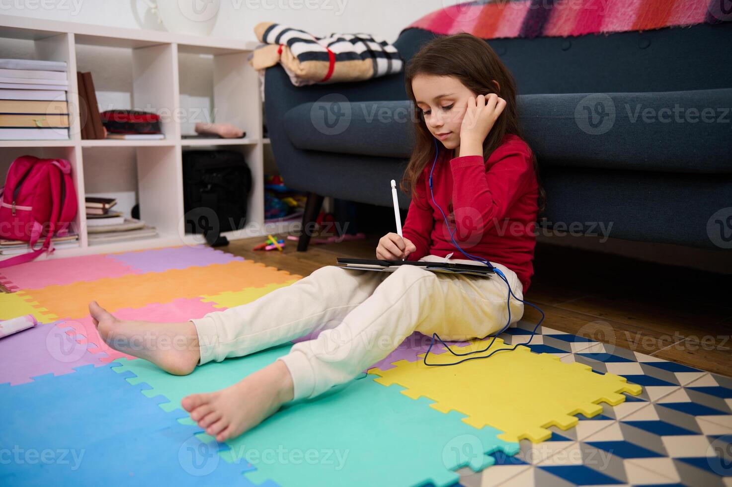 Adorable little child girl putting on earphones, sitting on a multicolored puzzle carpet, with a digital tablet, ready for online lesson. Distance education. Kid entertainment and development photo