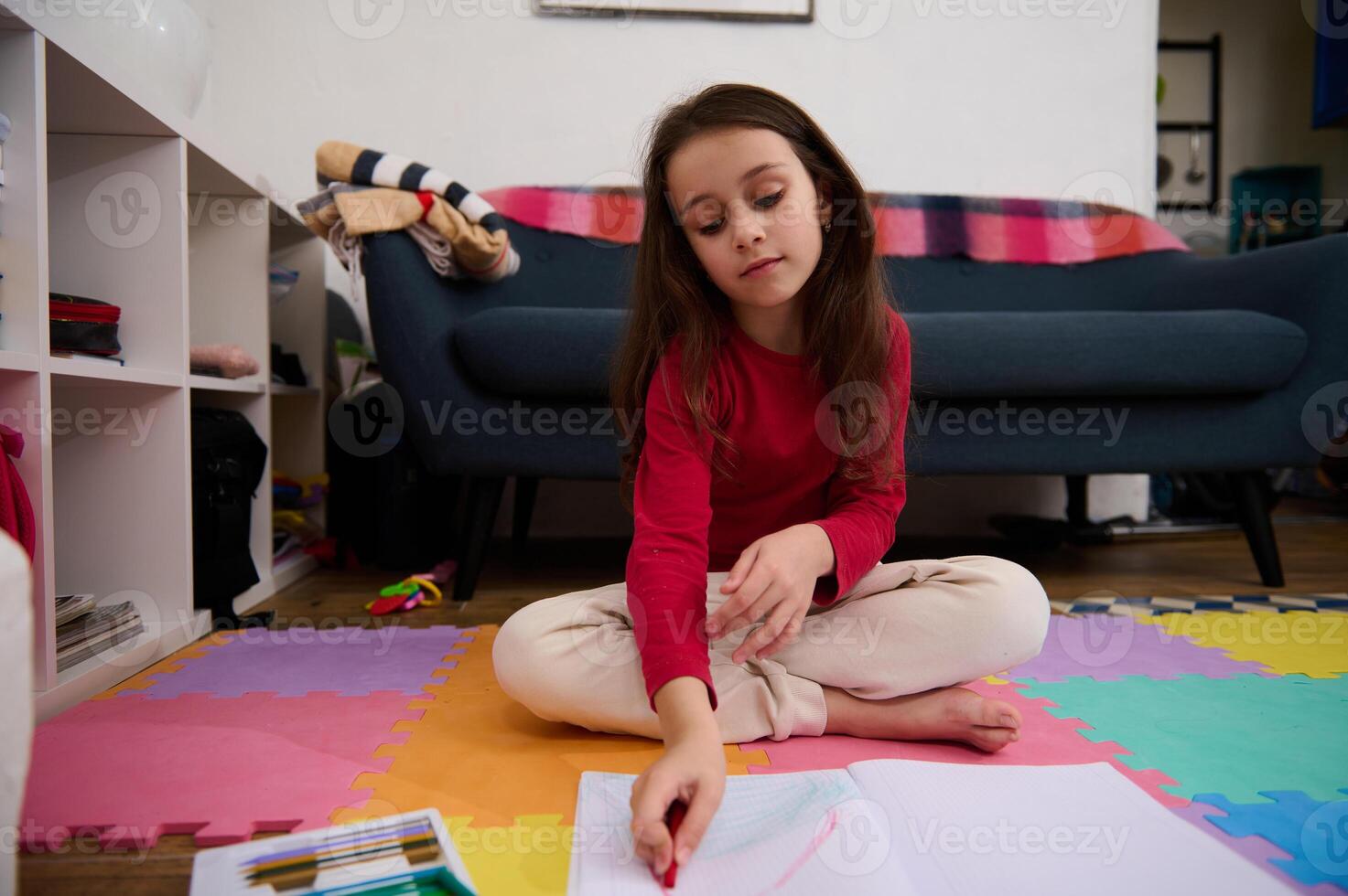 Little child girl with long hair, sitting on the puzzle carpet at home, doing homework, studying, drawing picture with colorful pencils. People, kids education and entertainment. Art and creativity photo