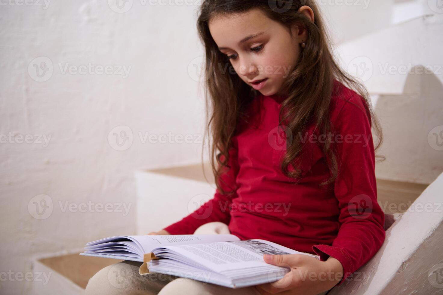 Authentic portrait of smart little kid girl leafing through the pages of a hardcover book, reading book, sitting on the steps at home, over white wall background of a rural house. Copy space for ads photo