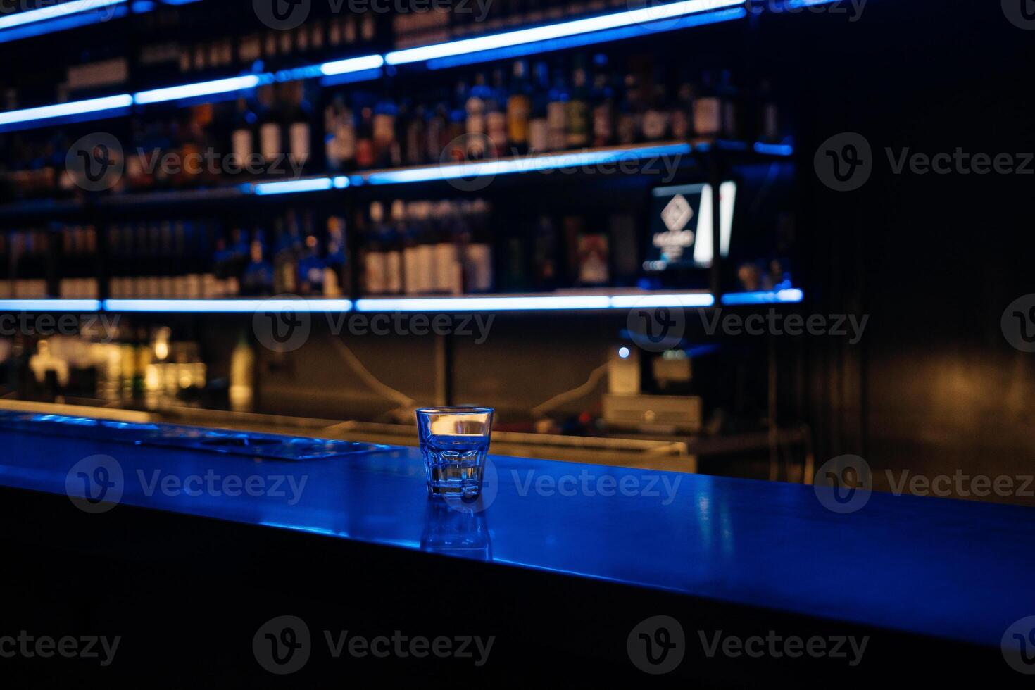 A bar with a blue counter and shelves of liquor. The bar is empty and the lights are on. photo
