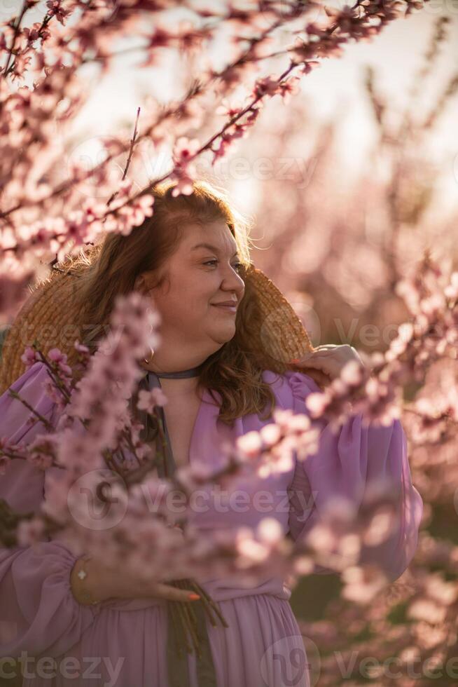 Woman blooming peach orchard. Against the backdrop of a picturesque peach orchard, a woman in a long pink dress and hat enjoys a peaceful walk in the park, surrounded by the beauty of nature. photo