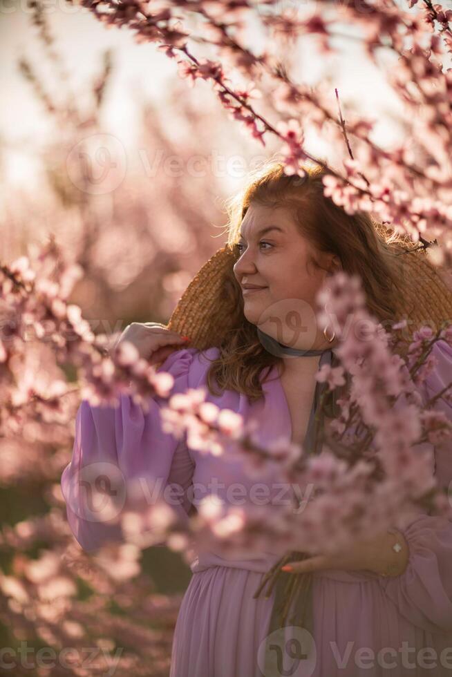 Woman blooming peach orchard. Against the backdrop of a picturesque peach orchard, a woman in a long pink dress and hat enjoys a peaceful walk in the park, surrounded by the beauty of nature. photo