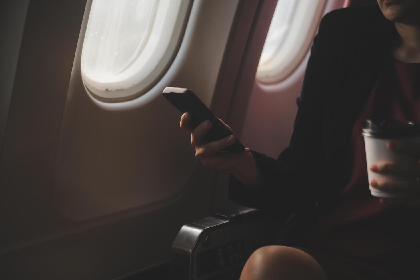 Blonde female tourist checking incoming notification on smartphone sitting on seat of airplane with netbook.Young businesswoman share media from telephone on laptop computer during plane flight photo