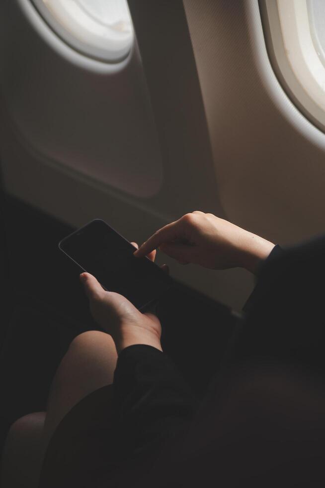Blonde female tourist checking incoming notification on smartphone sitting on seat of airplane with netbook.Young businesswoman share media from telephone on laptop computer during plane flight photo