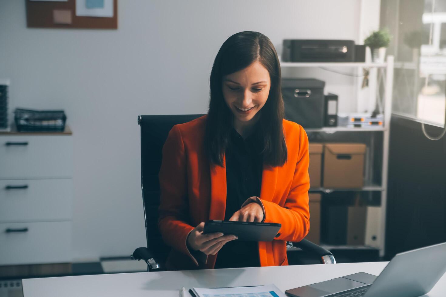 Smiling young Asian businesswoman working on a laptop computer at her desk in a bright modern home office, doing calculating expense financial report finance making notes on paper graph data document. photo