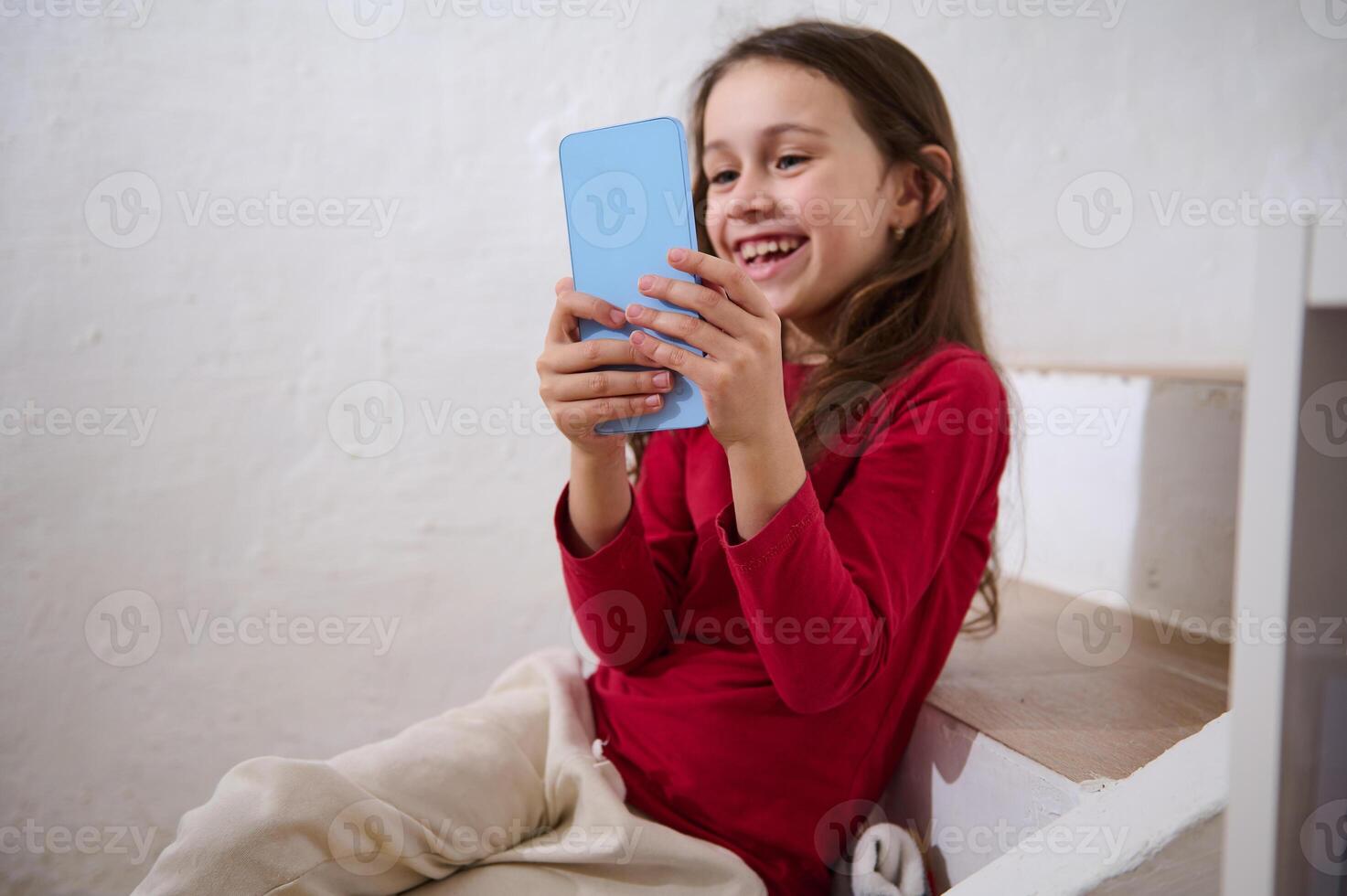 Focus on a modern blue smartphone in the hands of a blurred smiling little child girl, having fun watching , playing virtual games online. Digital addiction. Social media addicted kids photo
