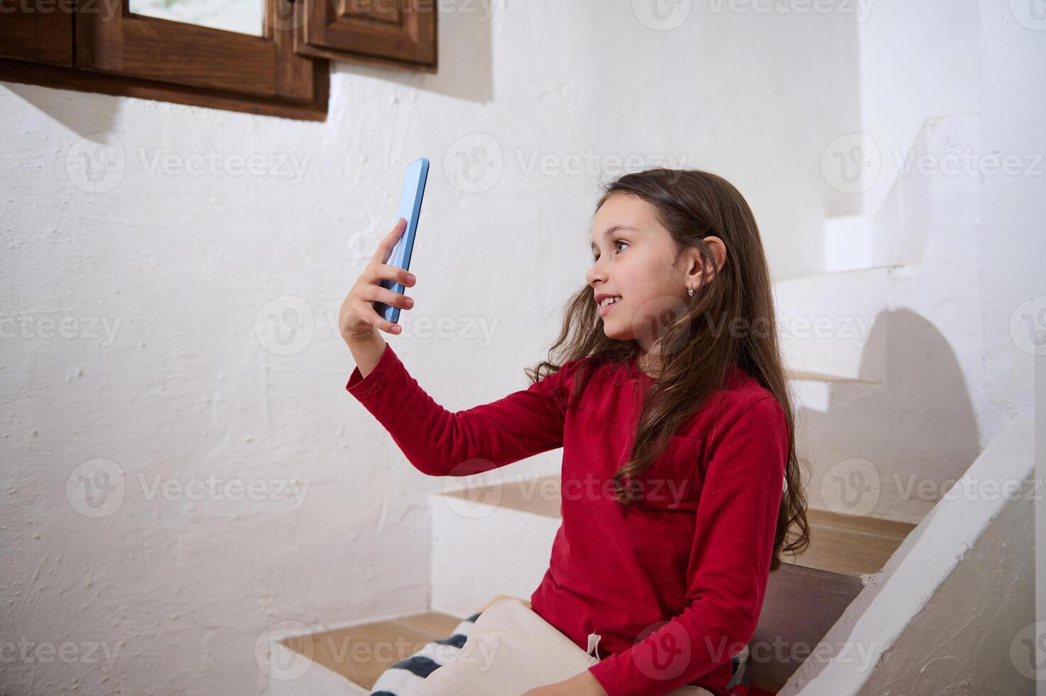 Beautiful Caucasian little child girl having fun on smart mobile phone, taking self-portrait selfie for sharing on social media, sitting on steps against white wall background in a rural house photo
