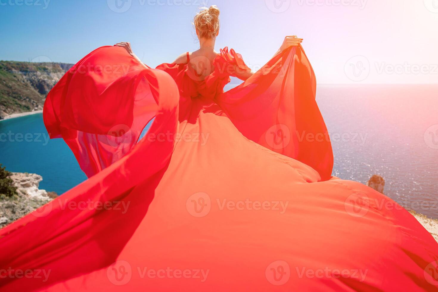 woman sea red dress. Blonde with long hair on a sunny seashore in a red flowing dress, back view, silk fabric waving in the wind. Against the backdrop of the blue sky and mountains on the seashore. photo