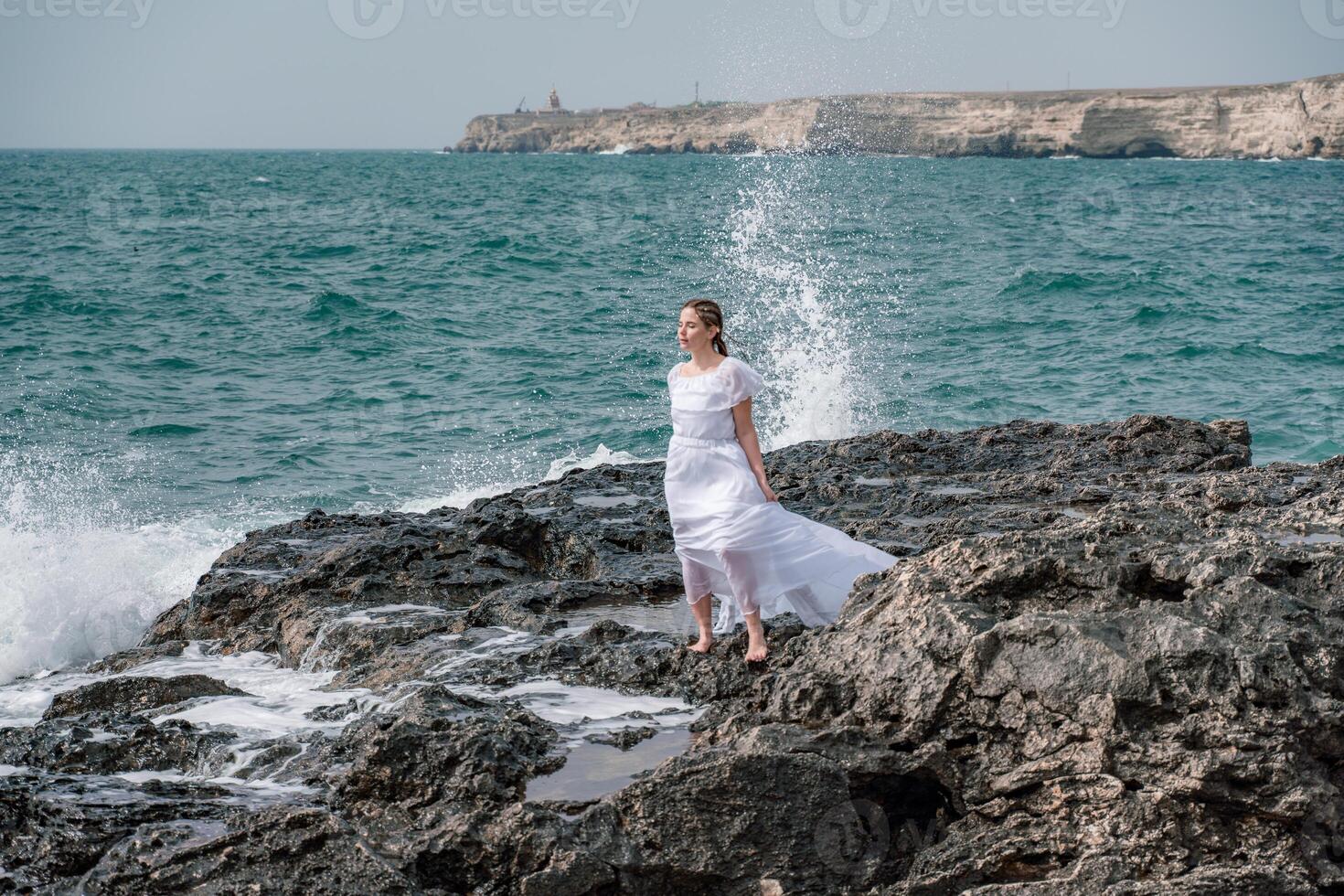 A woman stands on a rock in the sea during a storm. Dressed in a white long dress, the waves break on the rocks and white spray rises. photo