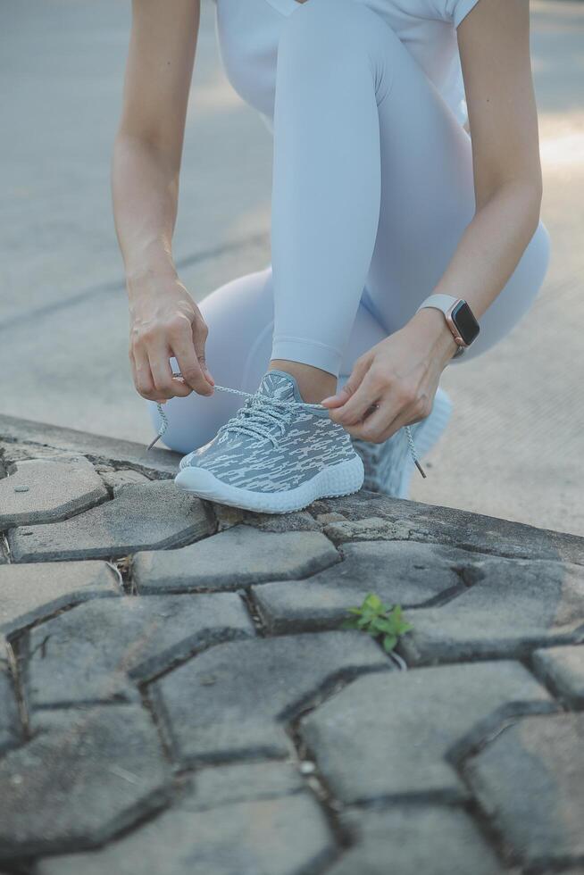 Mature fitness woman tie shoelaces on road. Cheerful runner sitting on floor on city streets with mobile and earphones wearing sport shoes. Active latin woman tying shoe lace before running. photo