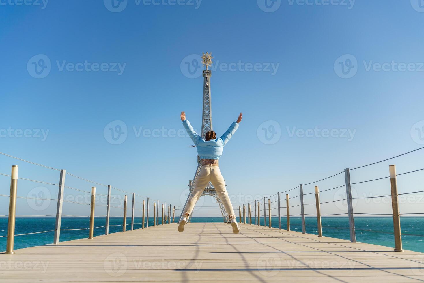Large model of the Eiffel Tower on the beach. A woman walks along the pier towards the tower, wearing a blue jacket and white jeans. photo