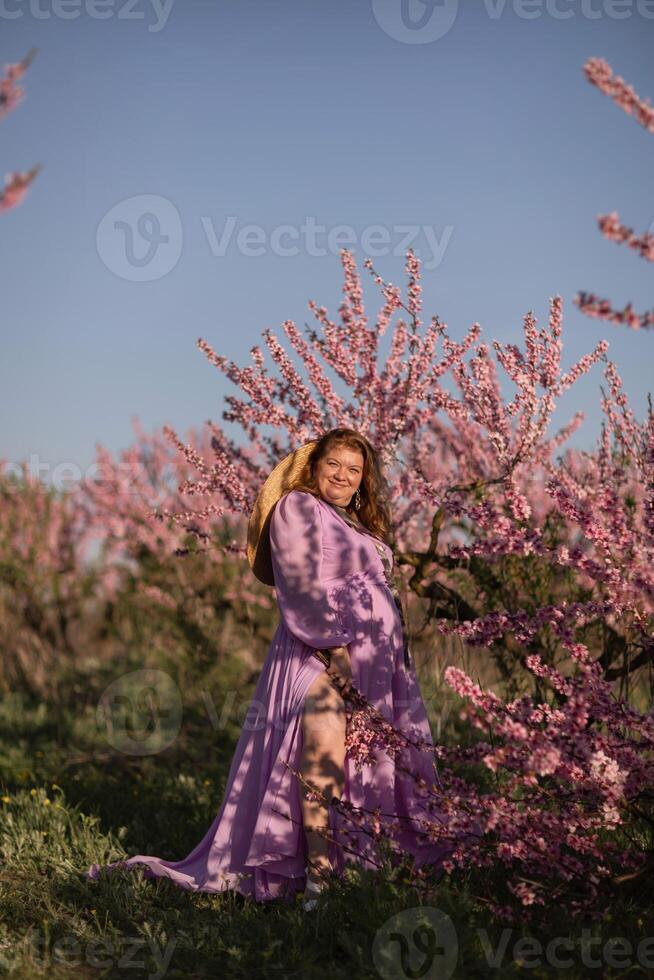 Woman blooming peach orchard. Against the backdrop of a picturesque peach orchard, a woman in a long pink dress and hat enjoys a peaceful walk in the park, surrounded by the beauty of nature. photo