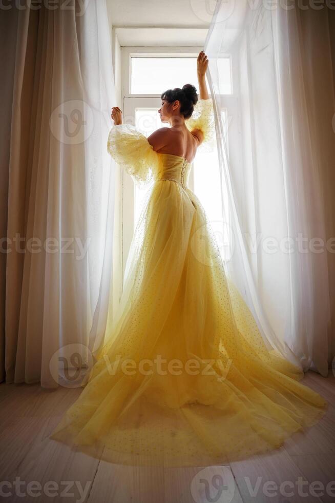 un mujer silueta en un dorado lujoso vestir en contra el antecedentes de un ventana sostiene un cortina con su manos. elegante dama en un amarillo largo seda vestir con desnudo atrás, espalda vista. foto