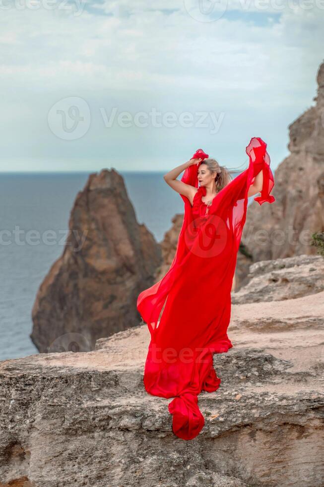 A woman in a red silk dress stands by the ocean, with mountains in the background, as her dress sways in the breeze. photo