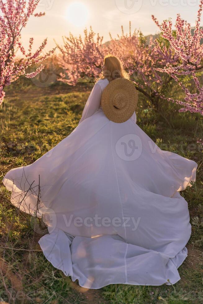 Woman blooming peach orchard. Against the backdrop of a picturesque peach orchard, a woman in a long white dress and hat enjoys a peaceful walk in the park, surrounded by the beauty of nature. photo