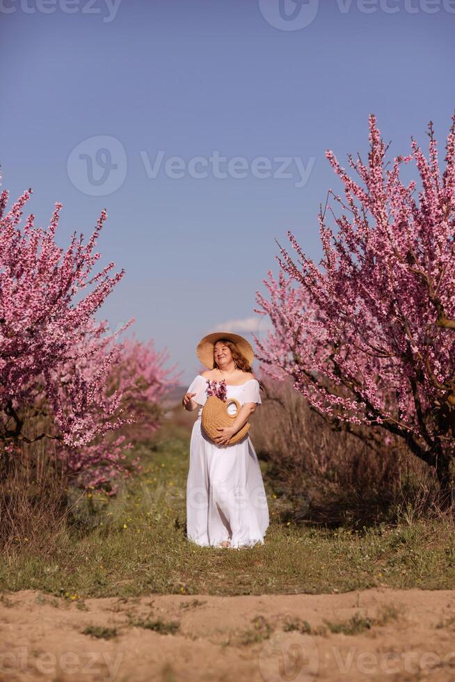 mujer floreciente melocotón huerta. en contra el fondo de un pintoresco melocotón huerta, un mujer en un largo blanco vestir y sombrero disfruta un pacífico caminar en el parque, rodeado por el belleza de naturaleza. foto