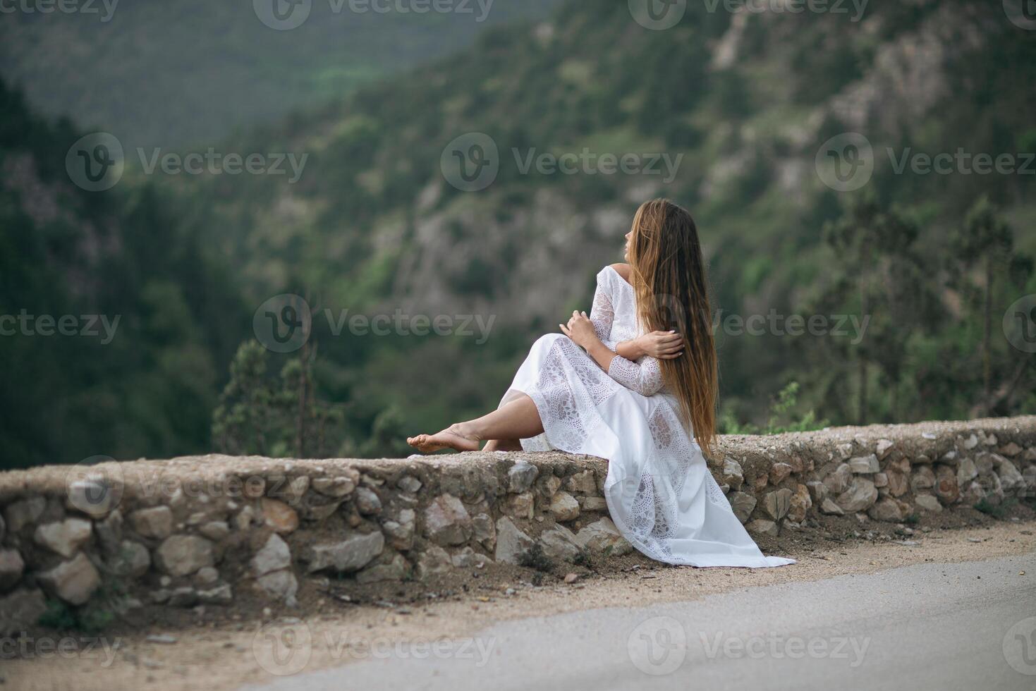 A woman in a white dress is sitting on a stone wall overlooking a mountain. The scene is serene and peaceful, with the woman's long hair blowing in the wind. Concept of calm and tranquility. photo