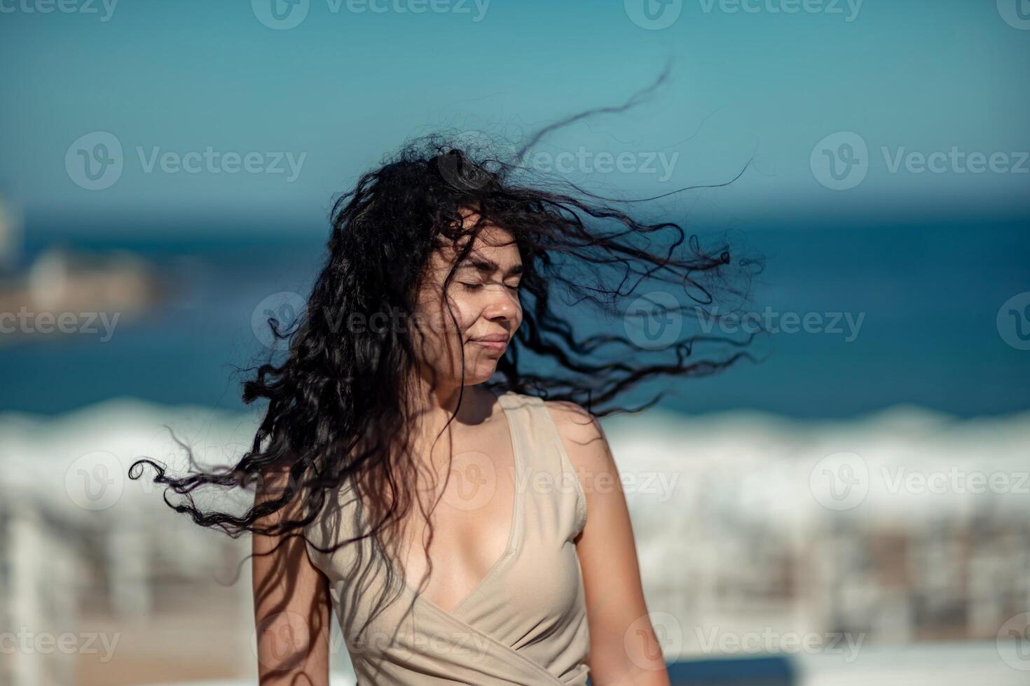 mar mujer descansar. retrato de un mujer con largo Rizado negro pelo en un beige vestir soportes en un balcón en contra el fondo de el mar. turista viaje a el mar. foto