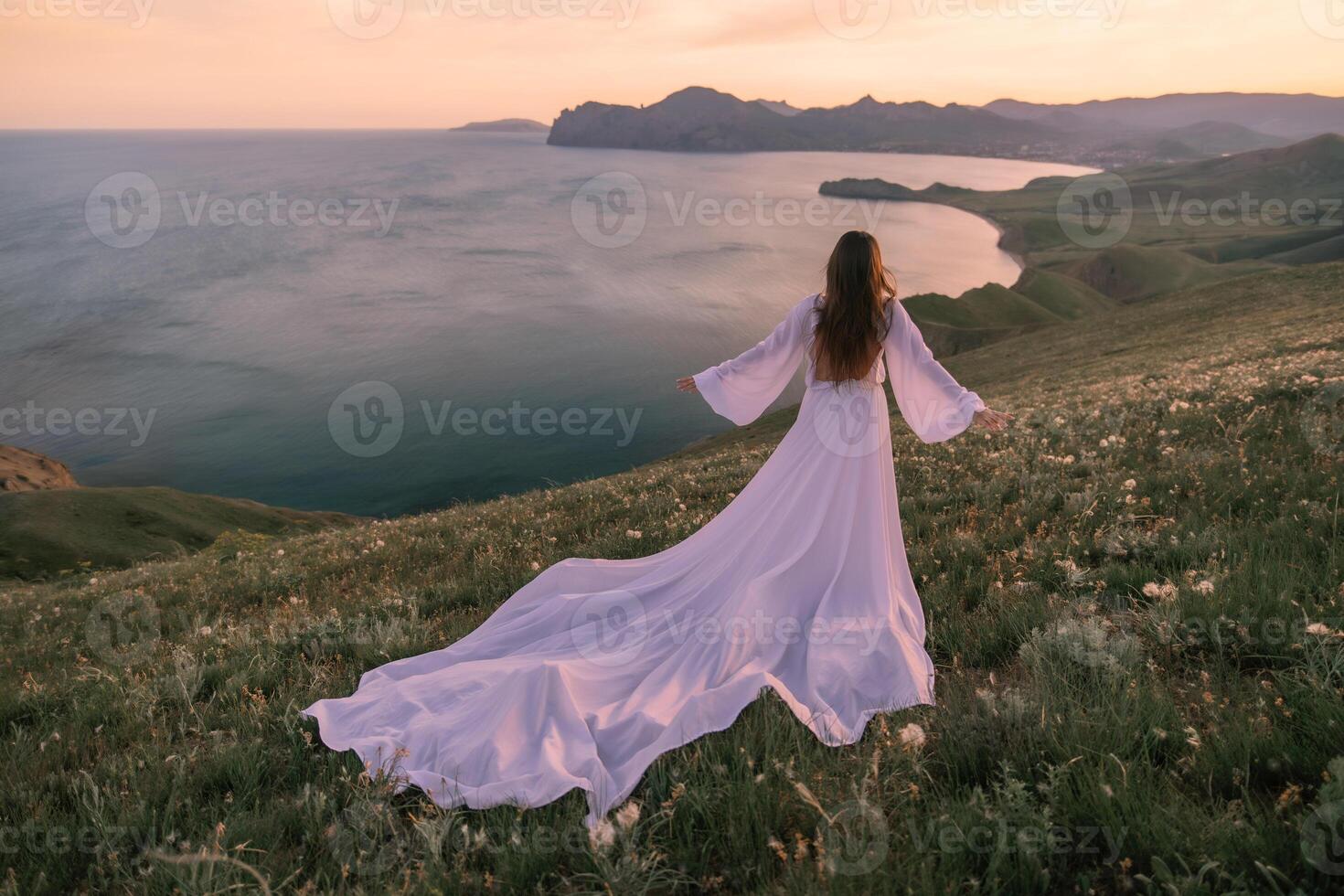A woman in a long white dress stands on a hill overlooking the ocean photo