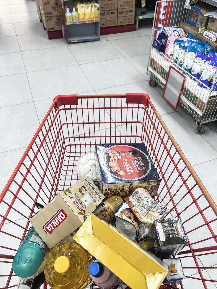 Shopping cart filled with food and drinks and supermarket shelves in the background, grocery shopping concept. Bekasi, Indonesia, April 22, 2024 photo