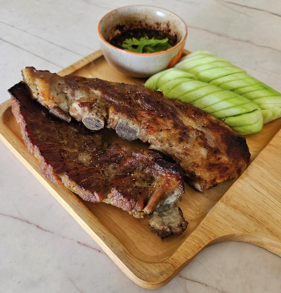 cerca arriba A la parrilla Cerdo costillas, rebanado Pepino y picante salsa en de madera bandeja. parilla comida con Fresco vegetales en blanco mármol. selectivo atención foto