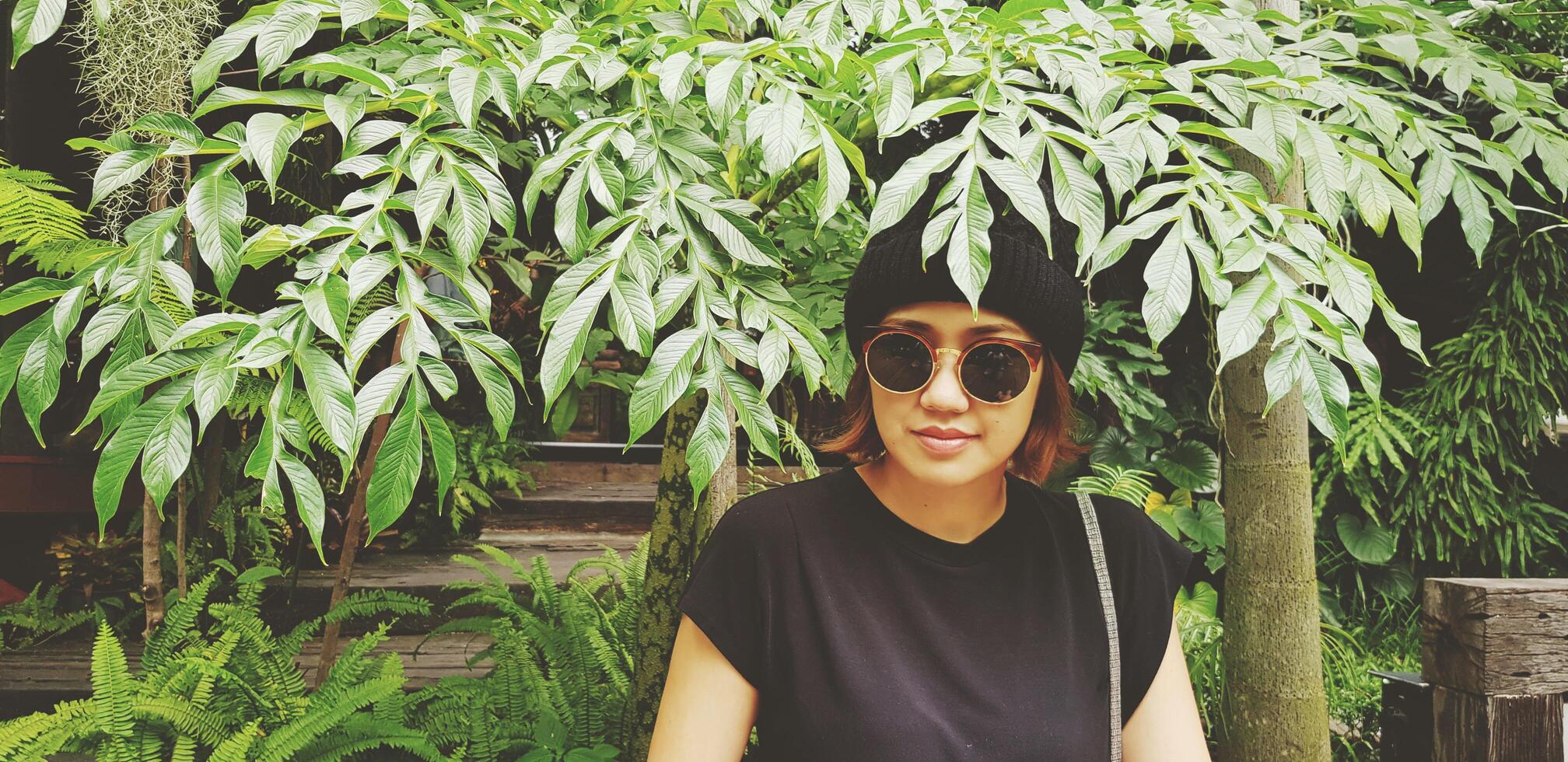 Asian woman is smiling, wearing sunglass and wool hat sitting on wooden bench under tree with green plant background. Girl in casual dress style in vintage tone color with copy space. photo