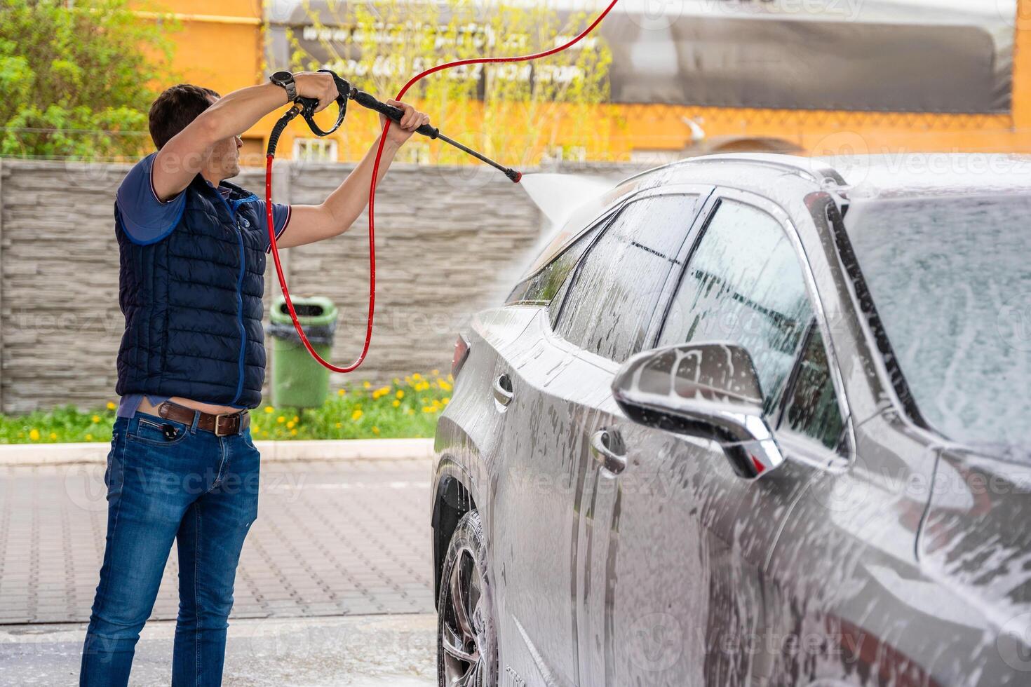 Young man washes his car at a self-service car wash using a hose with pressurized water and foam. photo