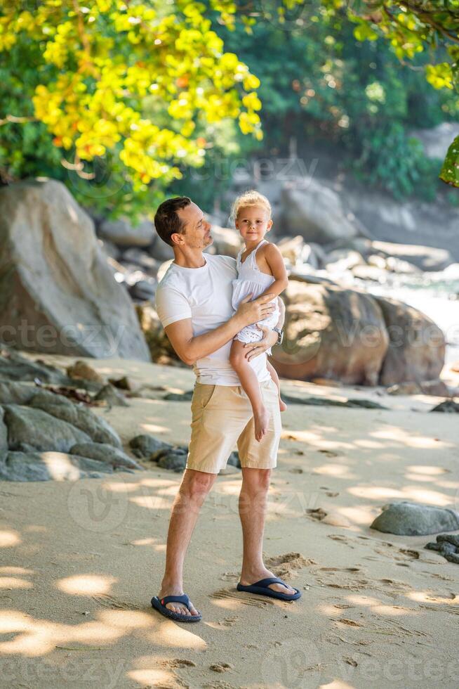 Little girl on father's hands smile and rejoice on seashore in the shade of trees and palms. Happy family concept photo