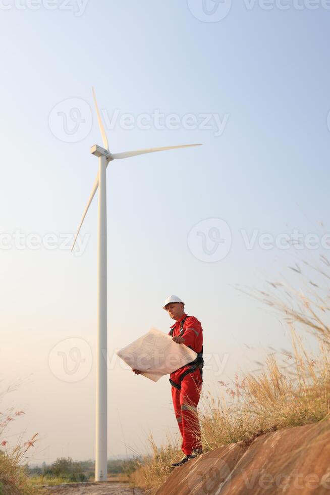 windmill engineer with red safety uniform hold drawing work at front of wind turbine farm photo