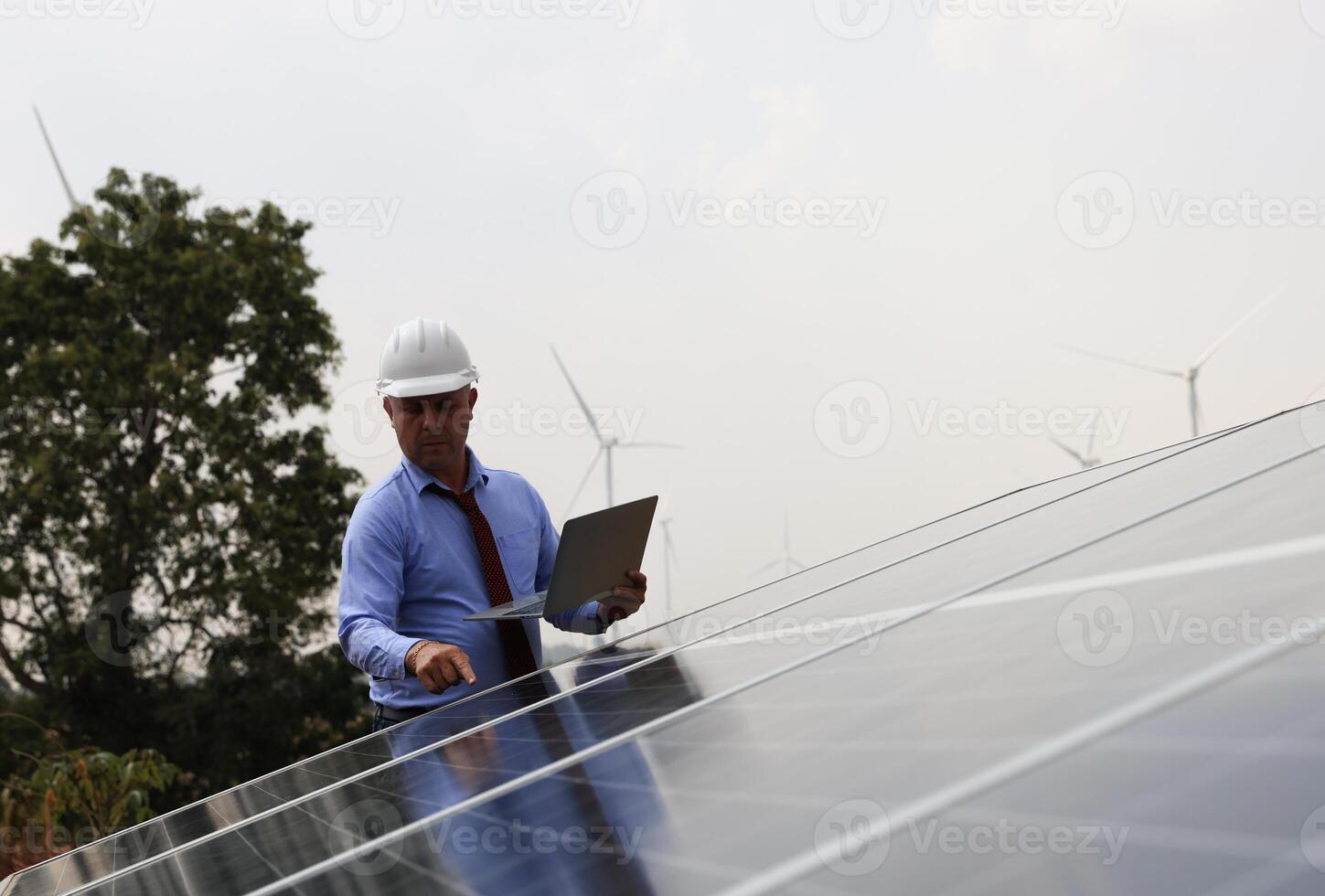 A senior male engineer inspects solar panels on the windmill farm. Clean energy. Engineers inspect a solar panel system with a turbine farm in the background, a renewable energy concept photo
