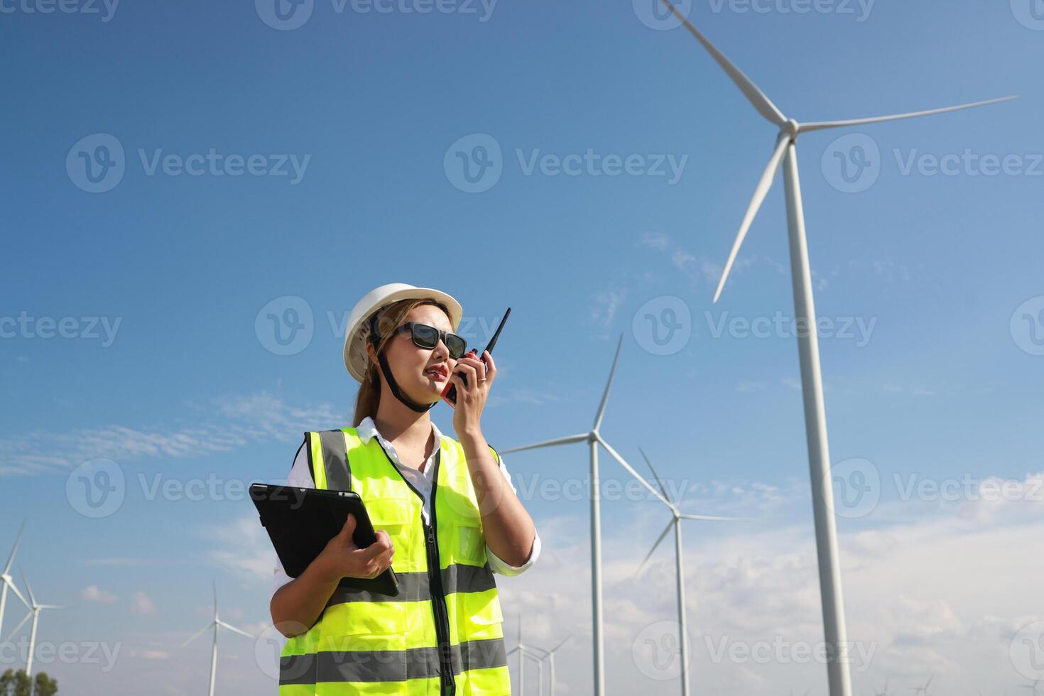 The confident woman in white helmet working with digital tablet and using walkie talkie at a renewable energy farm. Female inspector controlling the functioning of wind turbines outdoors photo