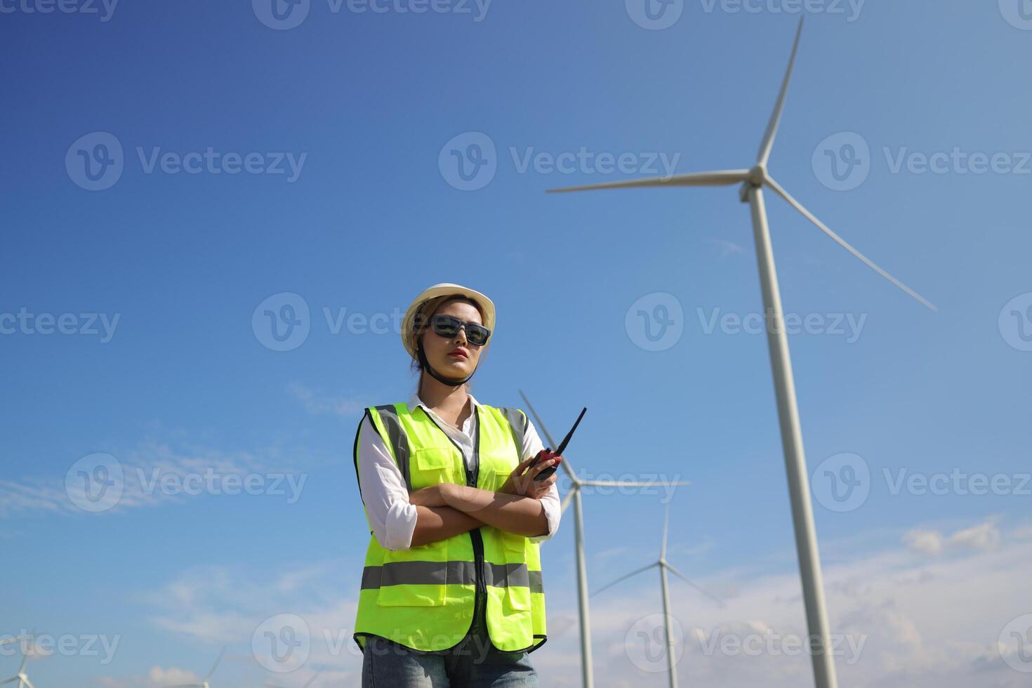 The confident woman in white helmet working with digital tablet and using walkie talkie at a renewable energy farm. Female inspector controlling the functioning of wind turbines outdoors photo