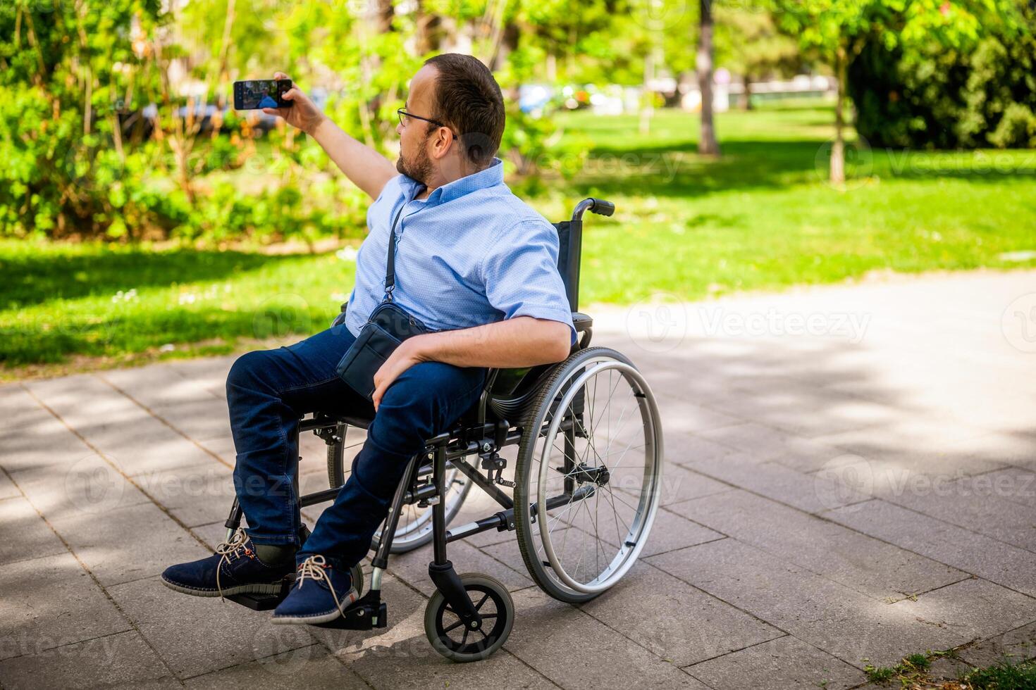 Portrait of happy man in wheelchair. He is enjoying sunny day in city park and taking selfie. photo