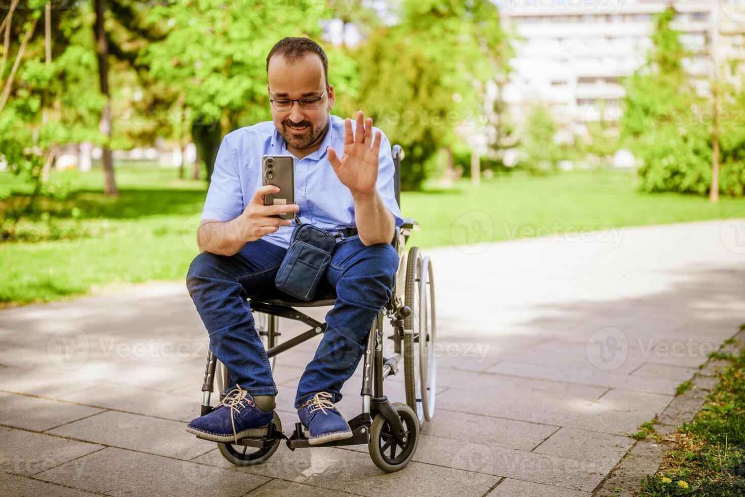 Portrait of happy man in wheelchair. He is enjoying sunny day in city park and messaging on smartphone. photo