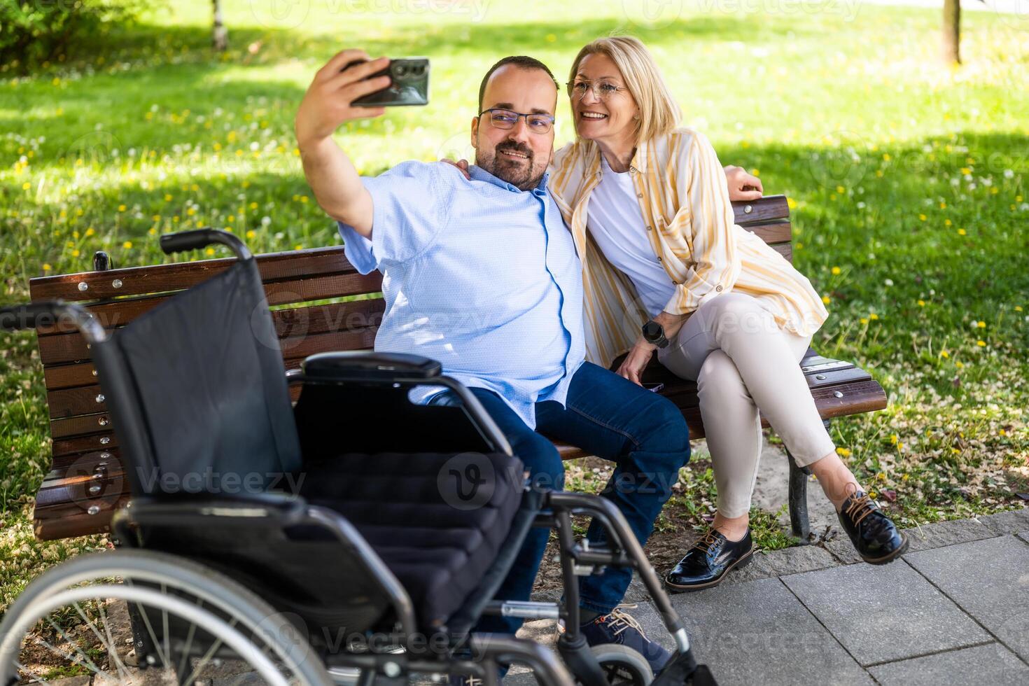 Man in wheelchair is spending time with his mother in park. They are taking selfie. photo