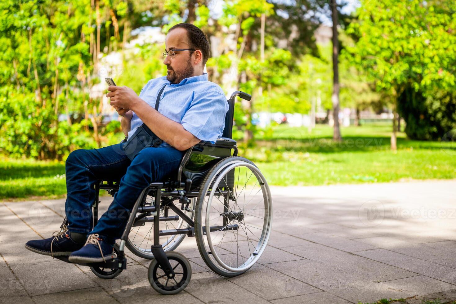 Portrait of man in wheelchair. He is enjoying sunny day in city park and messaging on smartphone. photo