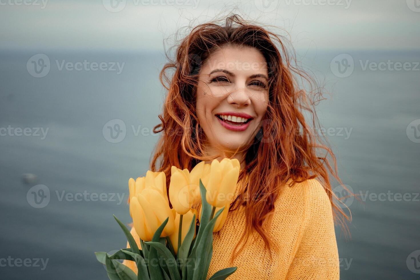 Portrait of a happy woman with hair flying in the wind against the backdrop of mountains and sea. Holding a bouquet of yellow tulips in her hands, wearing a yellow sweater photo