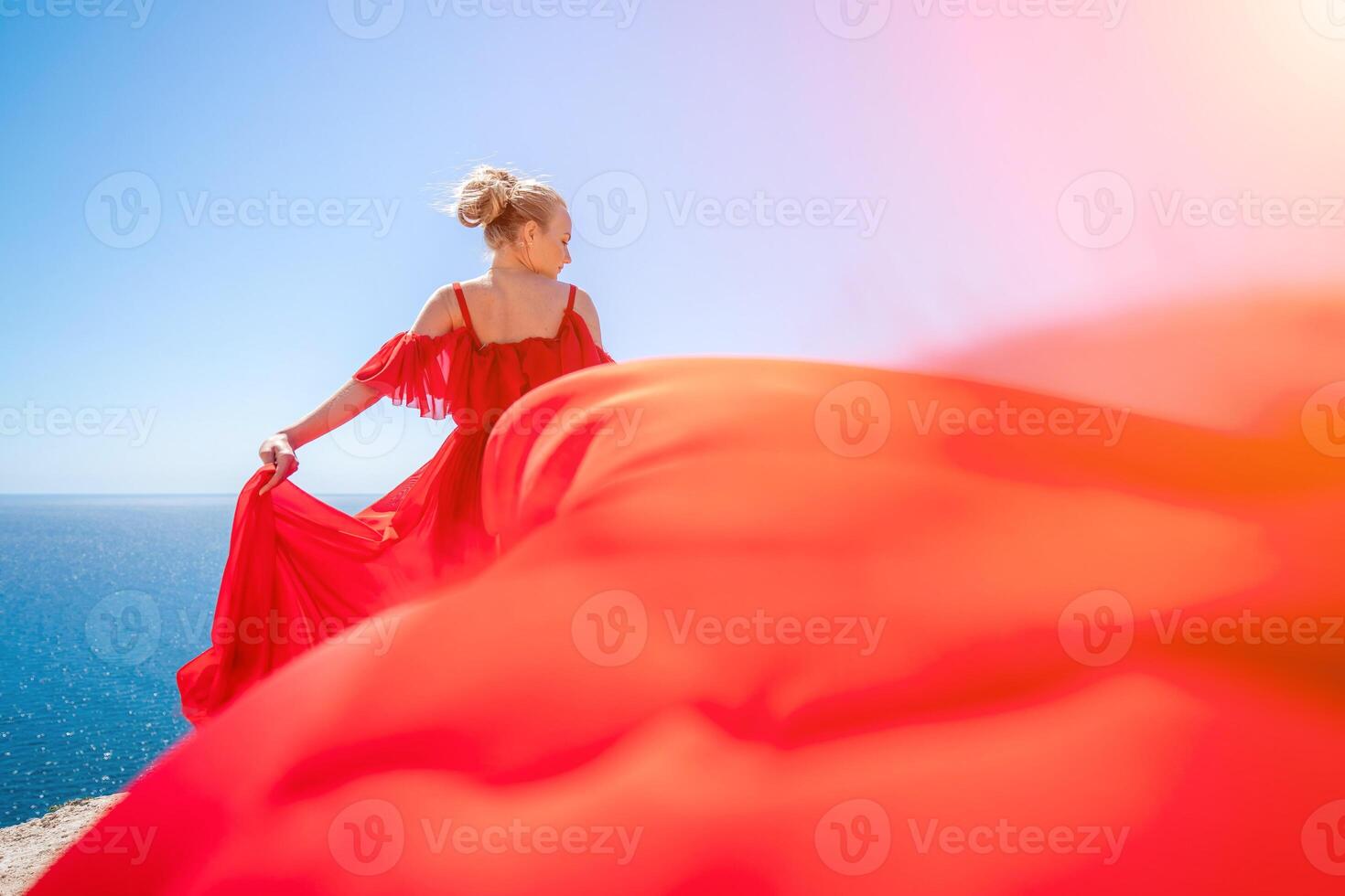 woman sea red dress. Blonde with long hair on a sunny seashore in a red flowing dress, back view, silk fabric waving in the wind. Against the backdrop of the blue sky and mountains on the seashore. photo