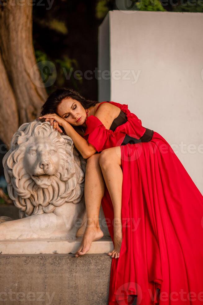 A woman in a red dress is sitting on a stone lion statue. The statue is white and has a lion's head. The woman is wearing a black belt and is looking at the camera. The scene has a calm. photo