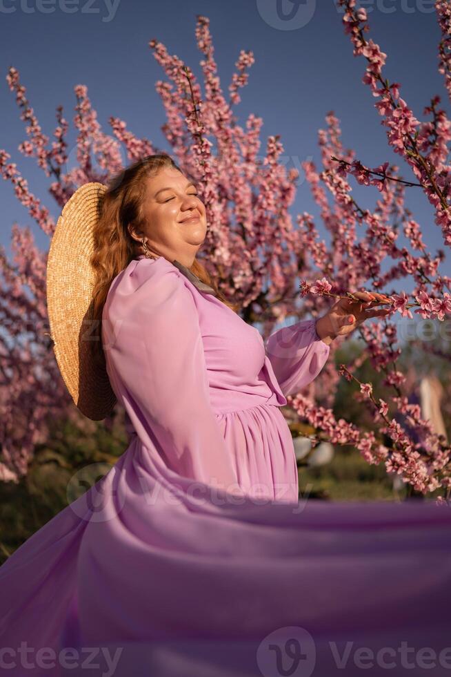 Woman blooming peach orchard. Against the backdrop of a picturesque peach orchard, a woman in a long pink dress and hat enjoys a peaceful walk in the park, surrounded by the beauty of nature. photo