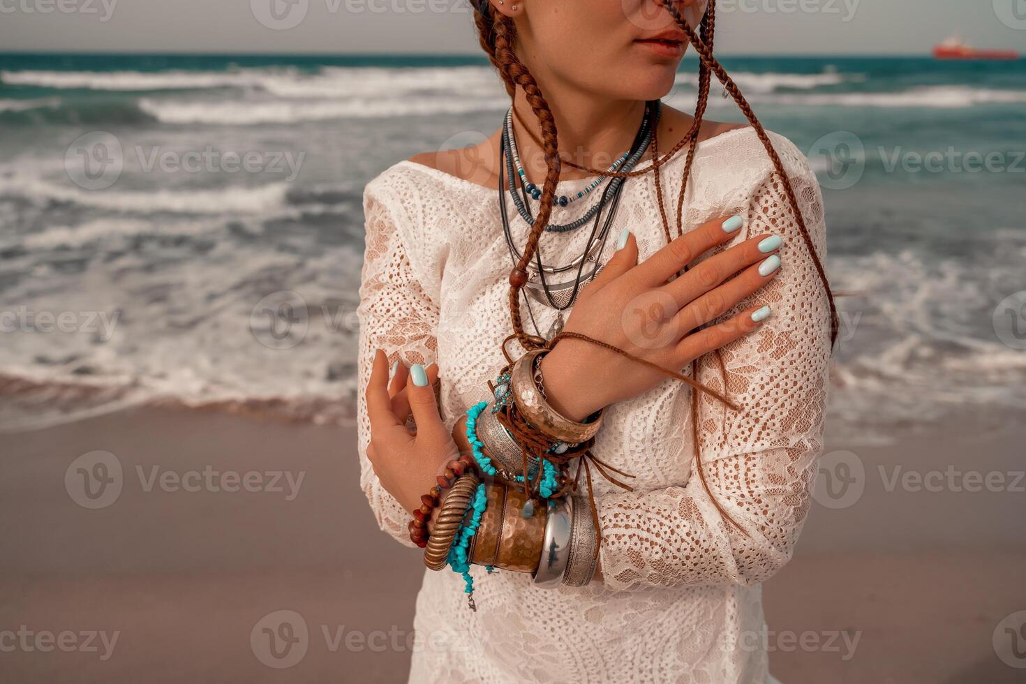 Model in boho style in a white long dress and silver jewelry on the beach. Her hair is braided, and there are many bracelets on her arms. photo