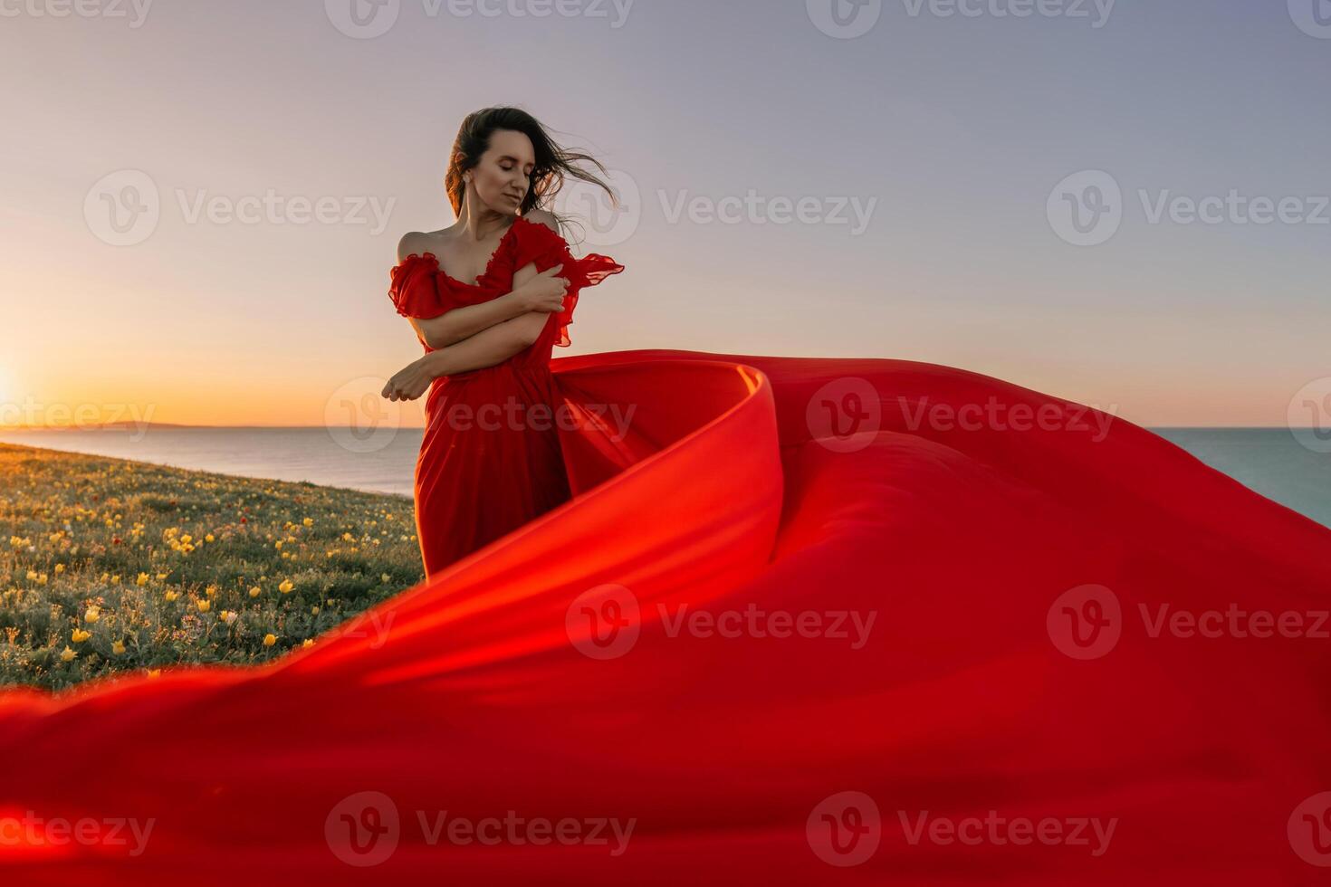 woman red dress standing grassy hillside. The sun is setting in the background, casting a warm glow over the scene. The woman is enjoying the beautiful view and the peaceful atmosphere. photo