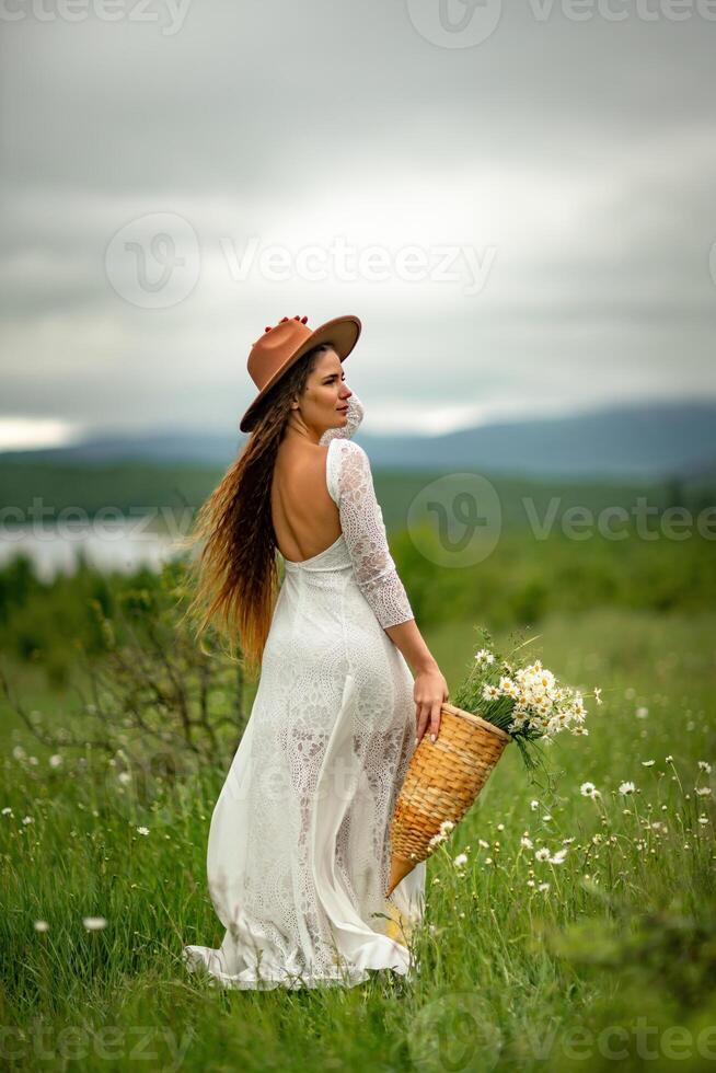 A middle-aged woman in a white dress and brown hat stands on a green field and holds a basket in her hands with a large bouquet of daisies. In the background there are mountains and a lake. photo