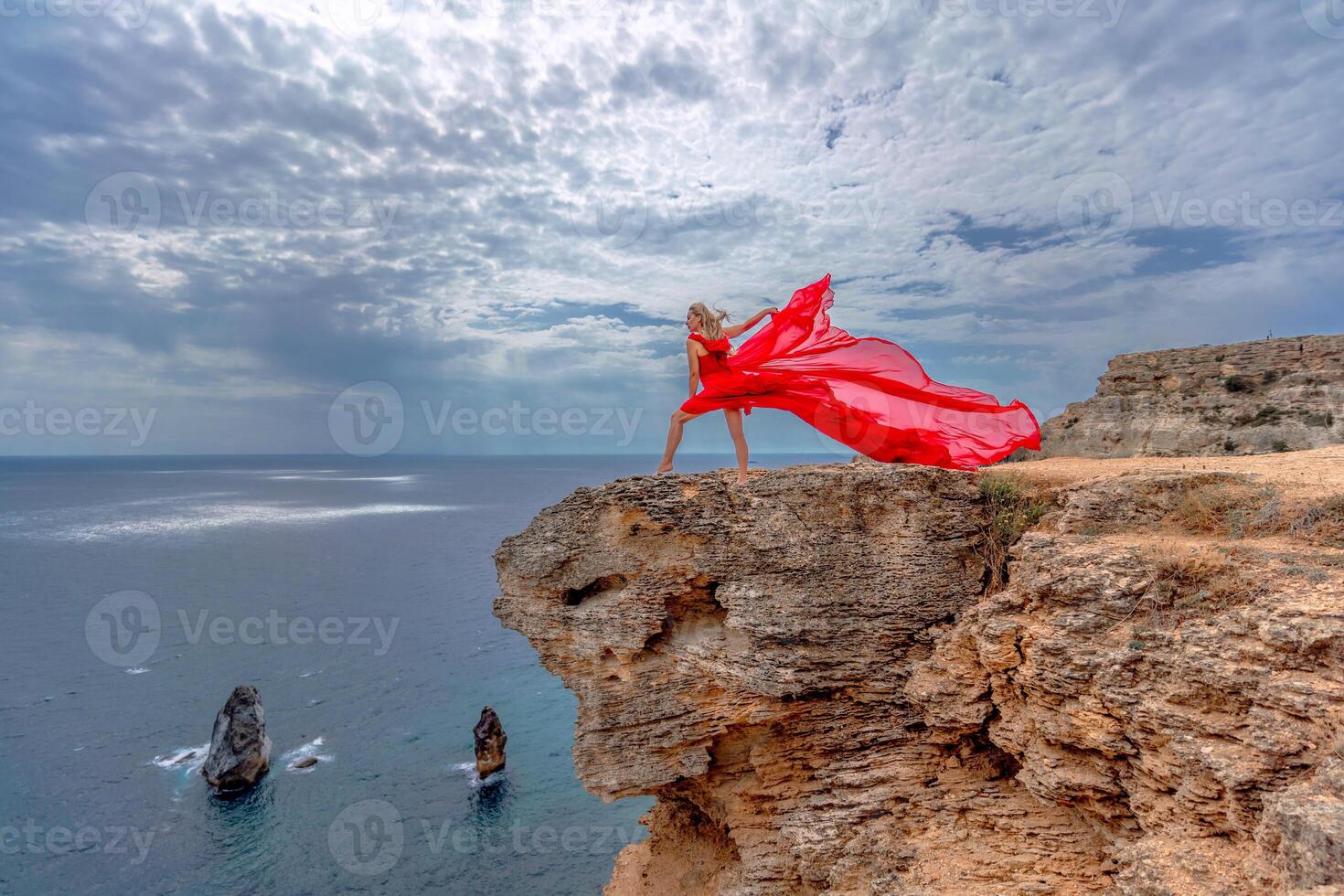 A woman in a red silk dress stands by the ocean, with mountains in the background, as her dress sways in the breeze. photo