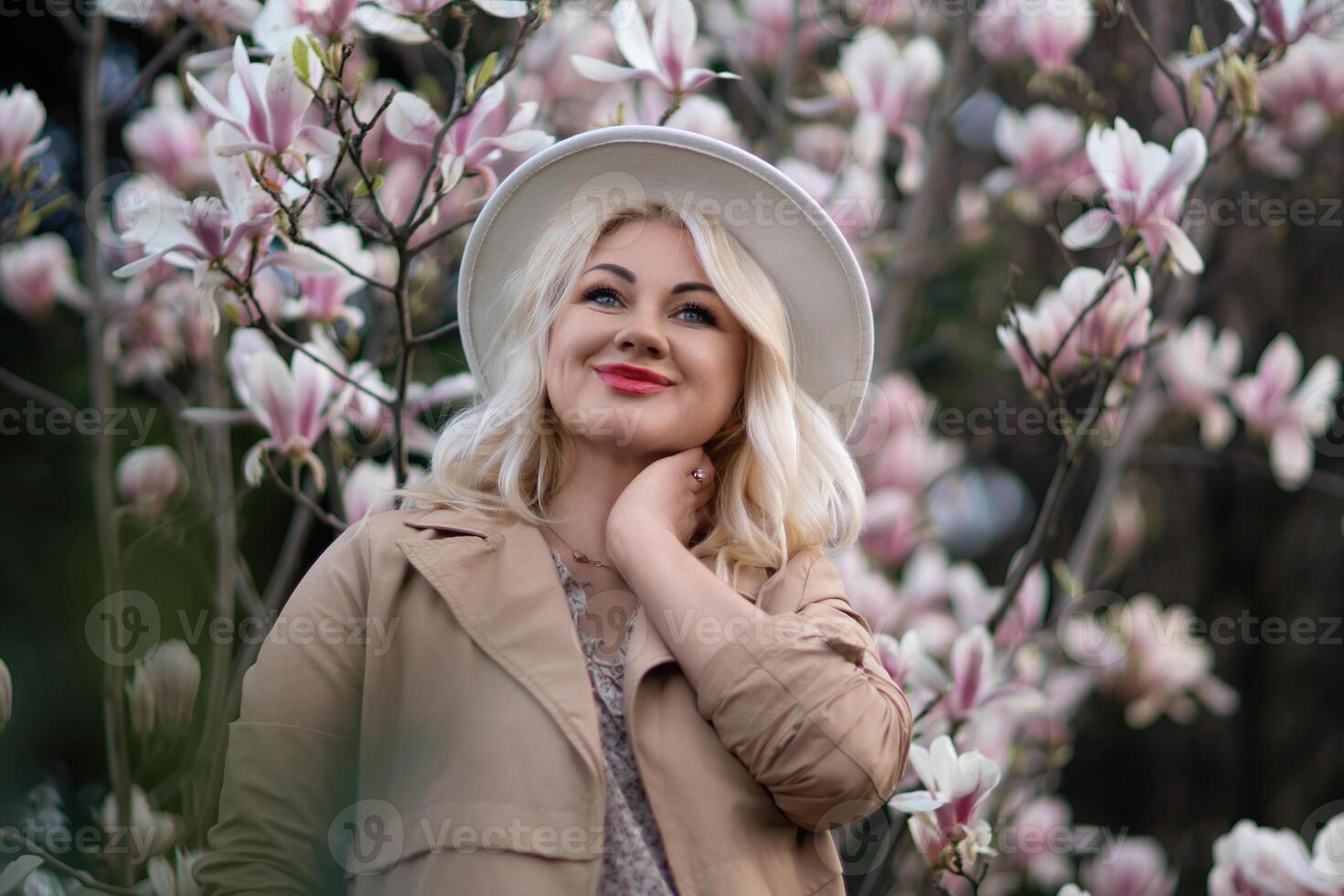 Magnolia flowers woman. A blonde woman wearing a hat stands in front of a tree with pink magnolia flowers. She is wearing a tan coat and a dress. photo