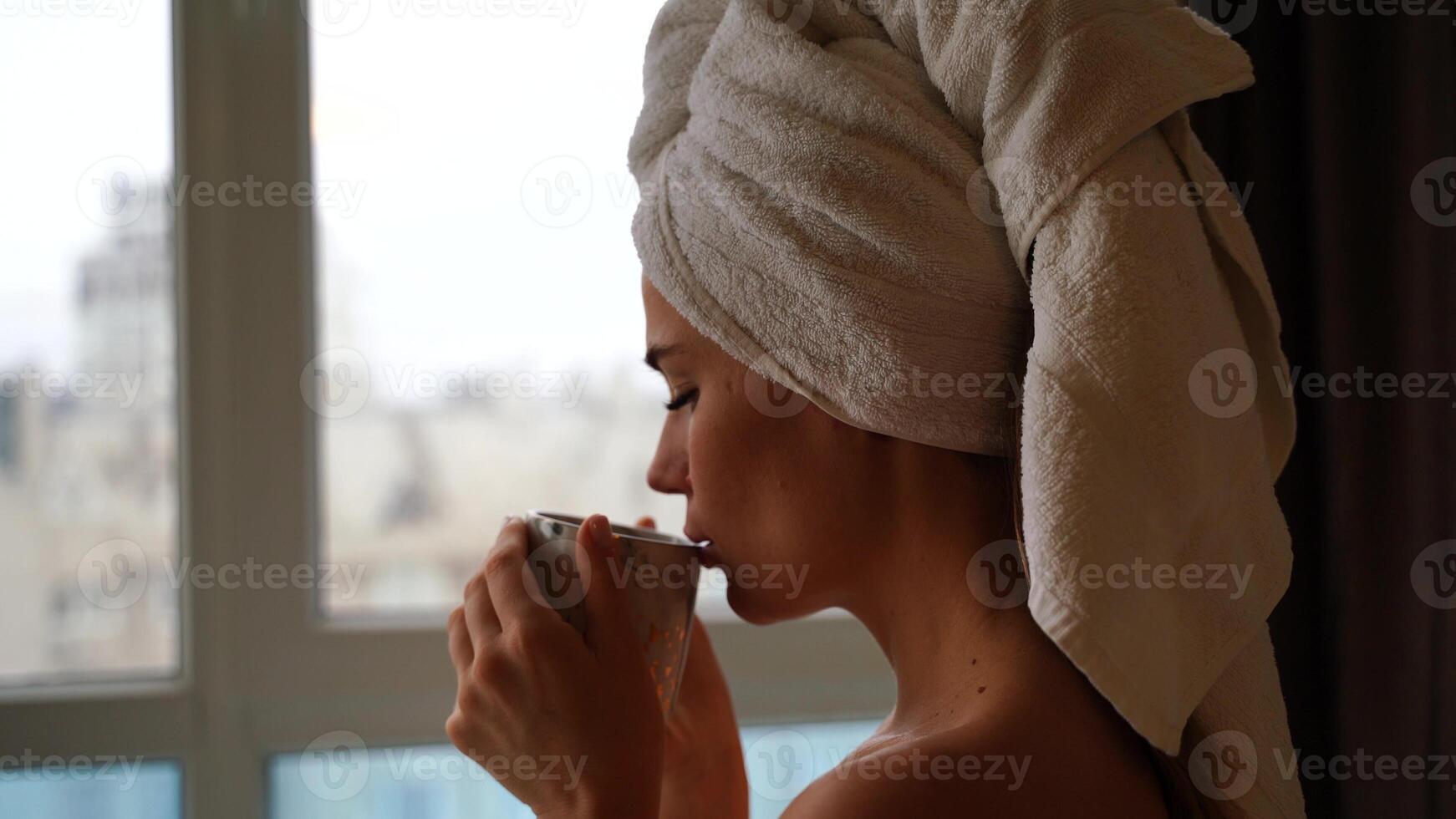 Middle aged woman looks good with bare shoulders in a white towel on her head holds a cup and drinks coffee or tea against the window photo