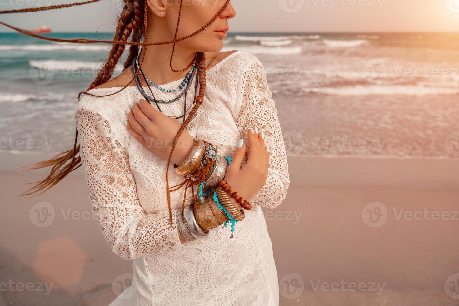 Model in boho style in a white long dress and silver jewelry on the beach. Her hair is braided, and there are many bracelets on her arms. photo