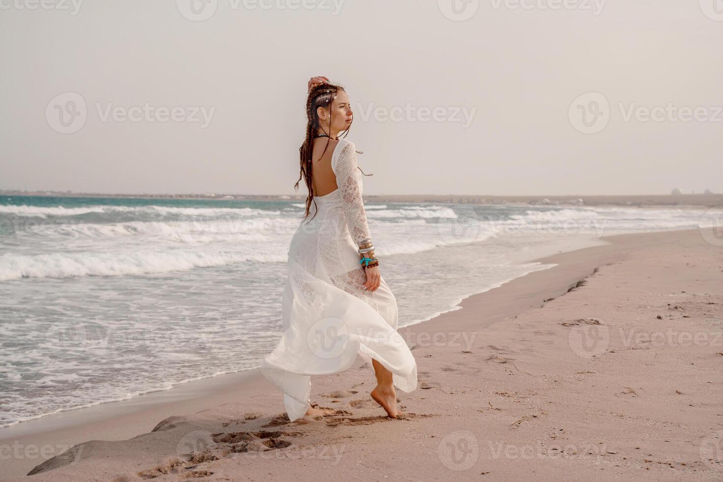 Model in boho style in a white long dress and silver jewelry on the beach. Her hair is braided, and there are many bracelets on her arms. photo